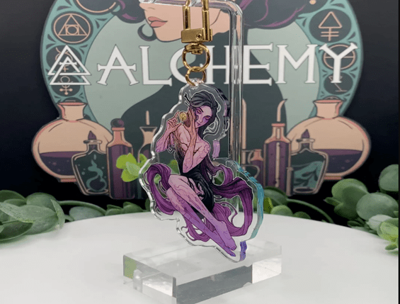 Custom acrylic and enamel pins displayed on stands in front of an illuminated Alchemy sign showcasing different pin styles and finishes